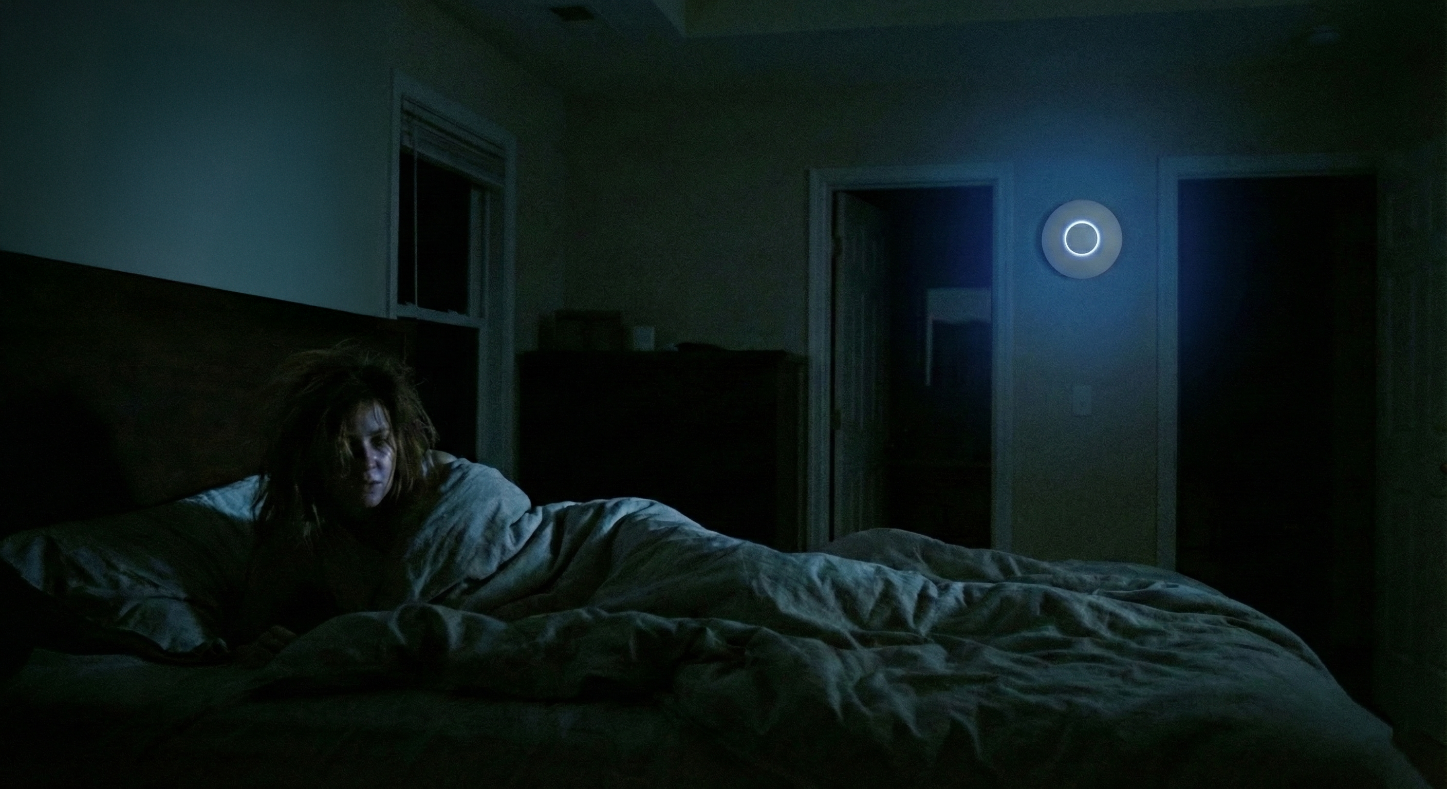 A restless woman lies awake in bed in the foreground, her face faintly illuminated by a cold blue cast. In the background, mounted on the wall near a doorway, a circular white UniFi access point glows with a bright blue LED ring. This device is the sole source of light, diffusing a soft, eerie blue ambiance across the room while the windows remain completely dark, creating an atmosphere of insomnia and technological intrusion.